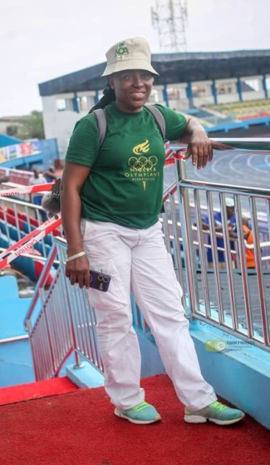A full-length shot of a Black woman standing outdoors at a sports stadium, smiling at the camera. She is wearing a green t-shirt with a gold logo that reads "Nigeria Olympians Association," white cargo pants, a light-colored bucket hat, and light blue sneakers. She is leaning against a stainless steel handrail next to a staircase with a red carpet runner, with an empty running track and stadium seating visible in the background