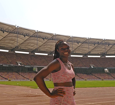 A medium shot of a smiling Black woman with long braids pulled back, standing confidently with hands on her hips on an outdoor running track. She is wearing a pink and white heathered sports bra and matching high-waisted shorts. In the background is a massive, empty sports stadium with rows of seating and a large white canopy roof under a bright sky.