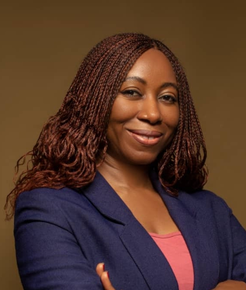 A woman in long braids smiling with hands akimbo. She is wearing a dark blue suit