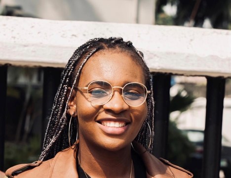 Headshot of a smiling woman with box braids, round glasses, and large hoop earrings wearing a brown jacket