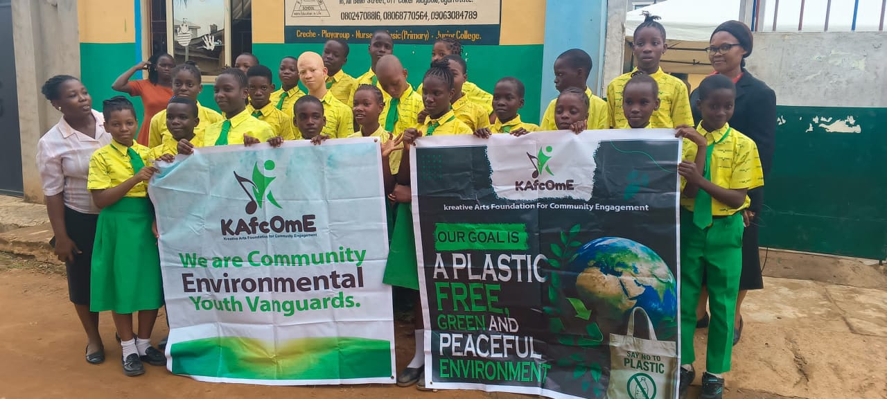 A group of school students in green-and-yellow uniforms pose outdoors alongside two instructors. Together, they hold two large banners for the Kreative Arts Foundation For Community Engagement (KAfComE). The banner on the left reads "We are Community Environmental Youth Vanguards." The banner on the right reads "OUR GOAL IS A PLASTIC FREE, GREEN AND PEACEFUL ENVIRONMENT" alongside illustrations of the Earth and a tote bag labeled "SAY NO TO PLASTIC."