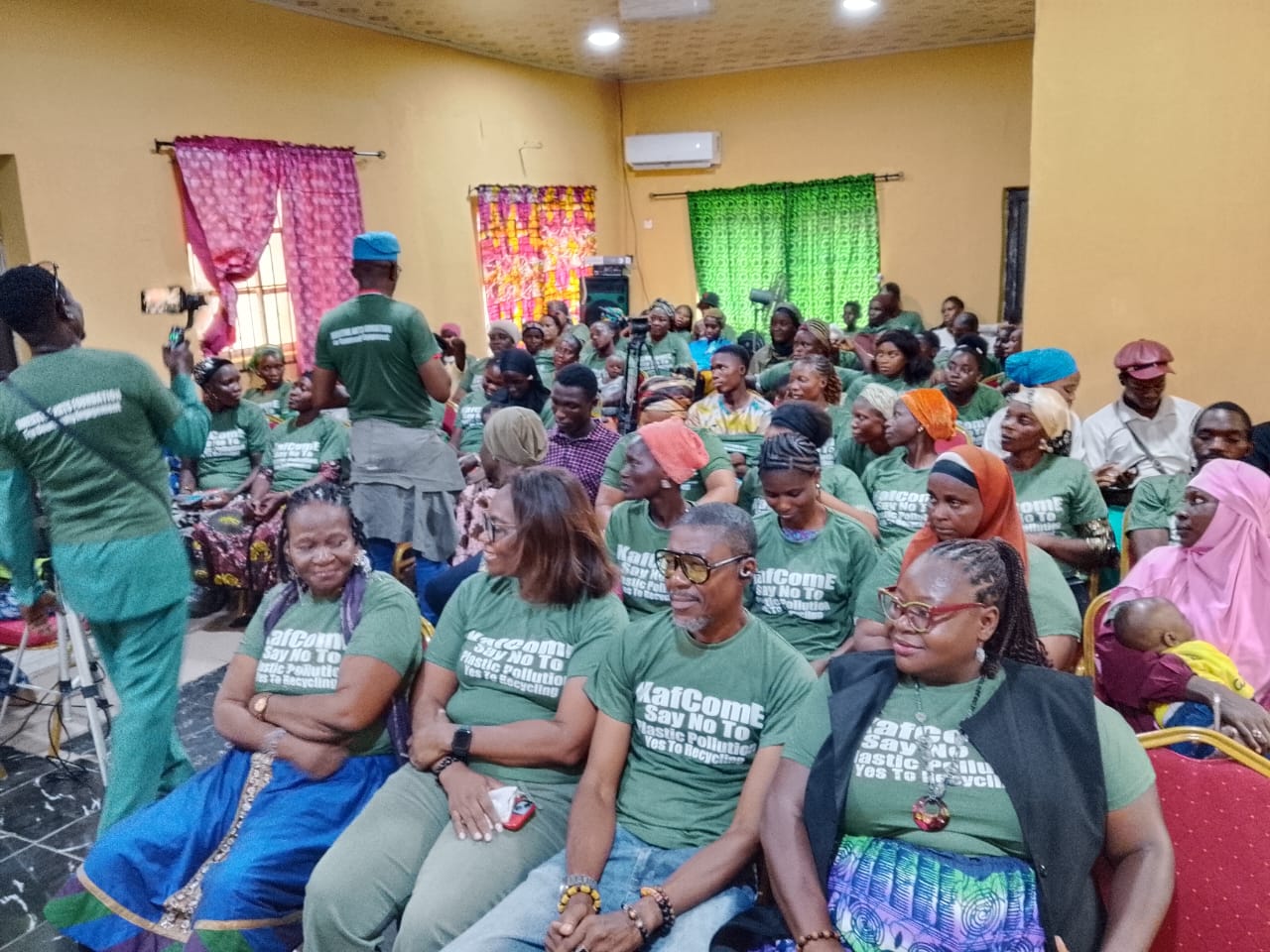 An indoor hall packed with adult participants listening attentively. Most people in the audience are wearing matching green t-shirts that read "KAfComE - Say No To Plastic Pollution, Yes To Recycling." On the far left, a videographer uses a gimbal stabilizer to film the seated crowd.