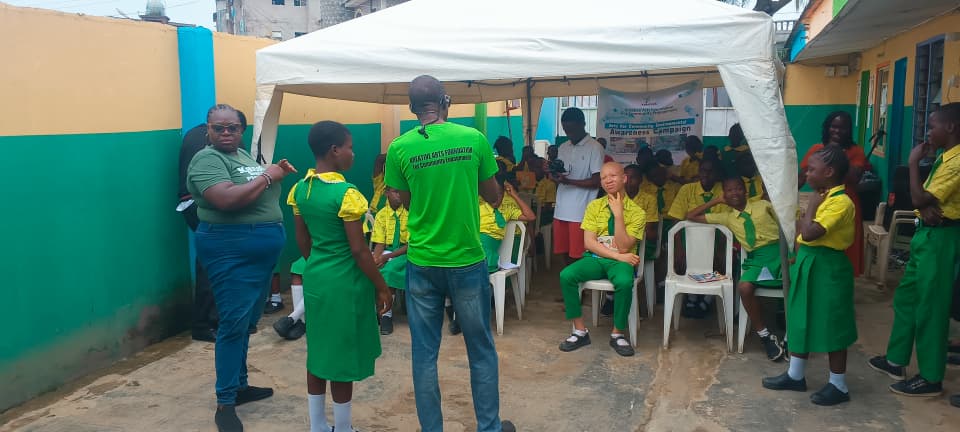 An outdoor workshop setup under a white pop-up canopy tent. Students wearing yellow shirts and green bottoms sit in white plastic chairs, listening to facilitators. In the center foreground, an instructor stands with his back to the camera, wearing a bright green shirt that reads "Kreative Arts Foundation For Community Engagement." To the left, another facilitator speaks to a student, while a campaign banner hangs in the background under the tent.