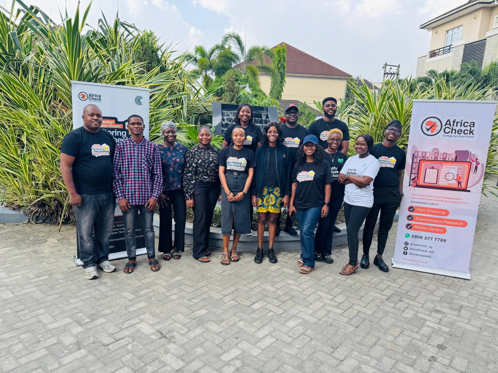 A group of twelve people stand smiling in a loose row outdoors on a paved stone courtyard. Tropical foliage, palm trees, and modern buildings with tiled roofs are visible in the background behind a small water fountain feature. The group is flanked by two white banner stands featuring the logo and branding for "Africa Check. Many of the participants are wearing matching black t-shirts bearing event graphics related to fact-checking and monitoring.