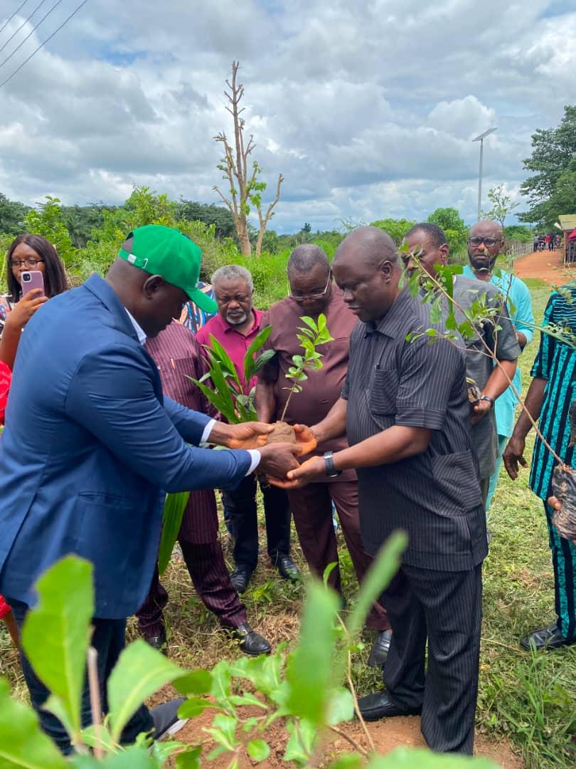 A man in a blue blazer and green cap handing a tree sapling to another man during an outdoor tree-planting ceremony surrounded by observers.
