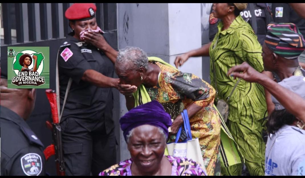 An outdoor photograph capturing a chaotic moment at a protest. On the left, a female Nigerian police officer in a black uniform and red beret covers her face with a patterned cloth while holding the hand of an elderly woman who is bent over, covering her mouth as if struggling from tear gas inhalation. In the lower foreground, a woman wearing a dark purple velvet headwrap looks toward the camera with a distressed expression. Other people are seen moving in the background, including a woman in a green patterned outfit and a man in a traditional striped cap. A green inset logo in the upper left corner depicts a map of Nigeria with a raised fist and text reading "End Bad Governance in Nigeria