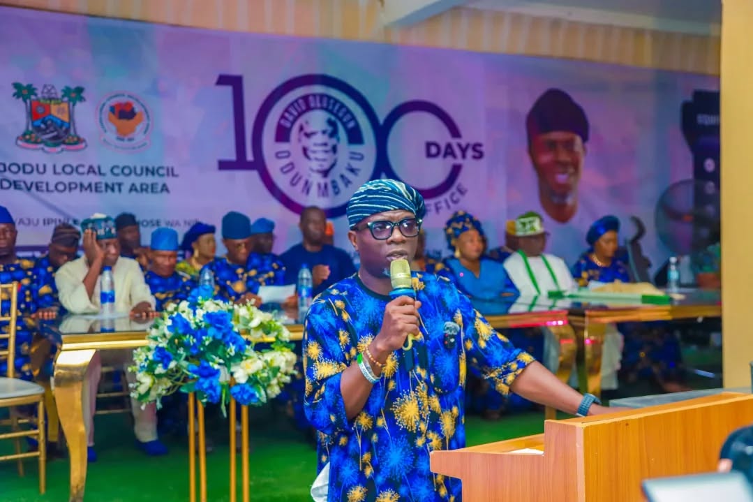A man wearing a bluish traditional attire, a cap and putting on shades holding a microphone in his left hand standing outdoors infront of a wooden podium. There are people seated at his back