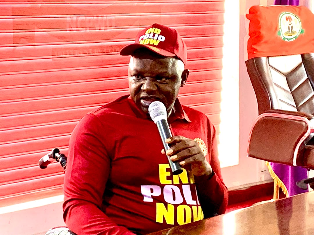 A medium shot of a man seated indoors, wearing a deep red long-sleeve shirt with yellow lettering reading "END POLIO NOW" and a matching red cap with similar text. He is holding a microphone and speaking, with a patterned red backdrop and a plush office chair visible behind him