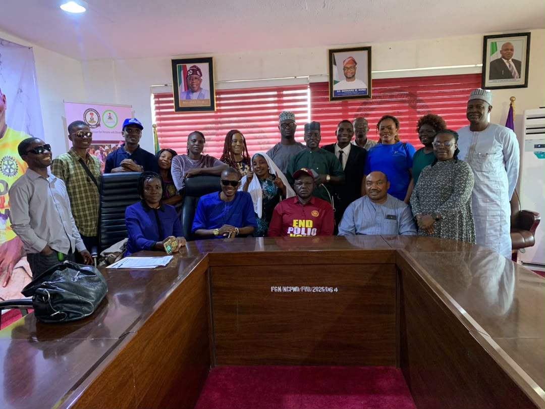 A wide-angle group photograph of multiple men and women standing and seated around a polished U-shaped conference table. The room features red window blinds, portraits hanging on the back wall, and bright overhead lighting. Some participants are smiling towards the camera, while a few wear sunglasses or traditional attire